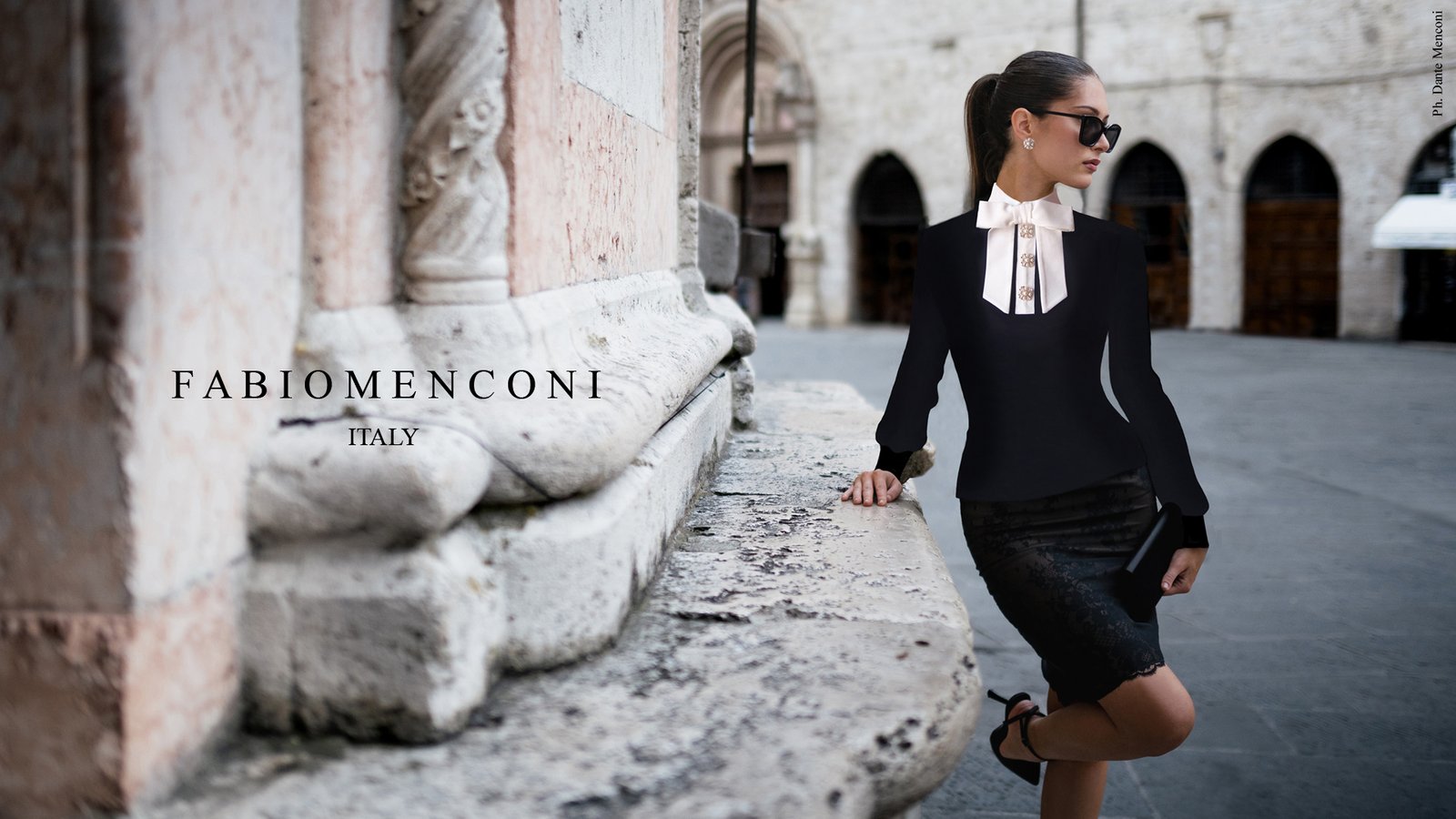 A model in a black jacket, white bow blouse and black pencil skirt leans on a carved stone base in a Perugia square, beside the name Fabio Menconi Italy.