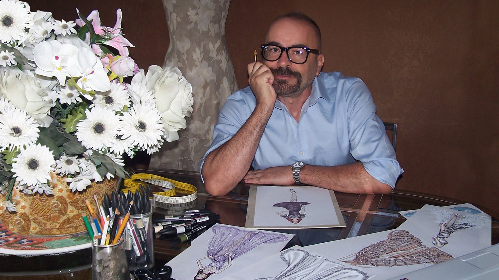 Fabio Menconi at his desk among his fashion sketches, with flowers and coloured pencils.