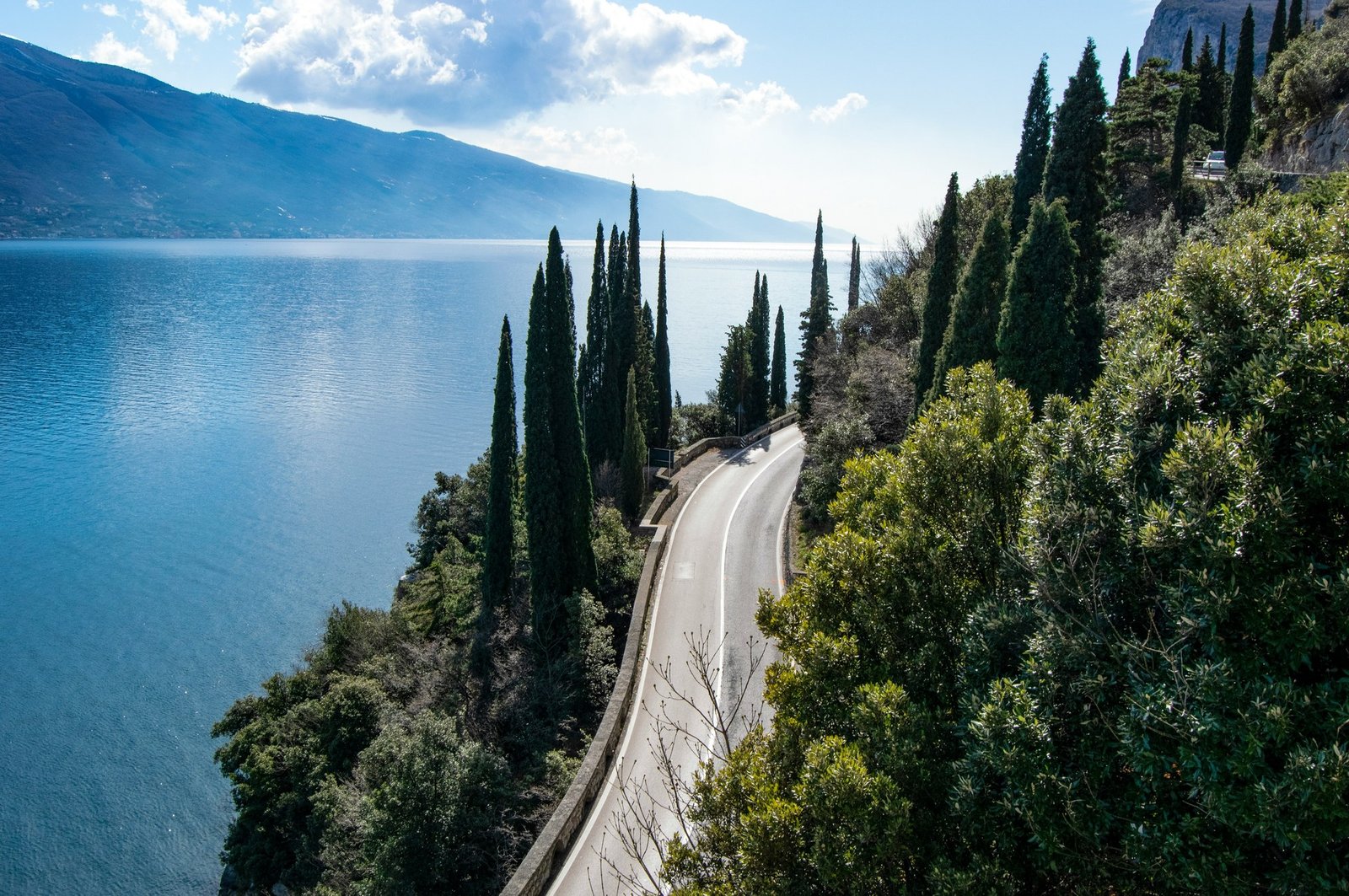 A road lined with cypress trees running along the shore of Lake Garda, with mountains across the water.