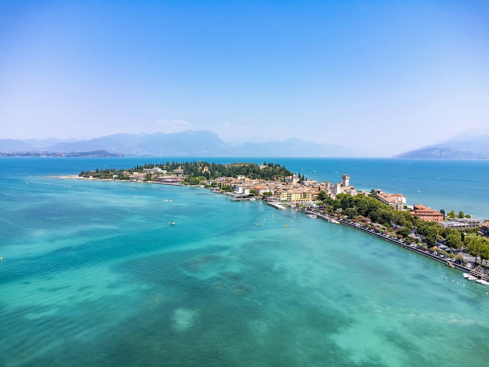Aerial view of a town on a narrow peninsula reaching into the turquoise water of Lake Garda.